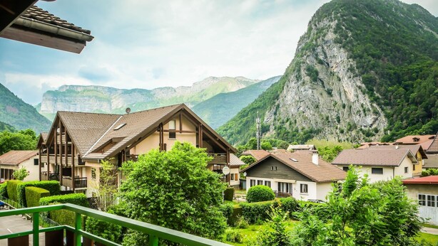 Un village de montagne aux maisons de style chalet traditionnel, entouré d’une végétation luxuriante et dominé par une falaise imposante.