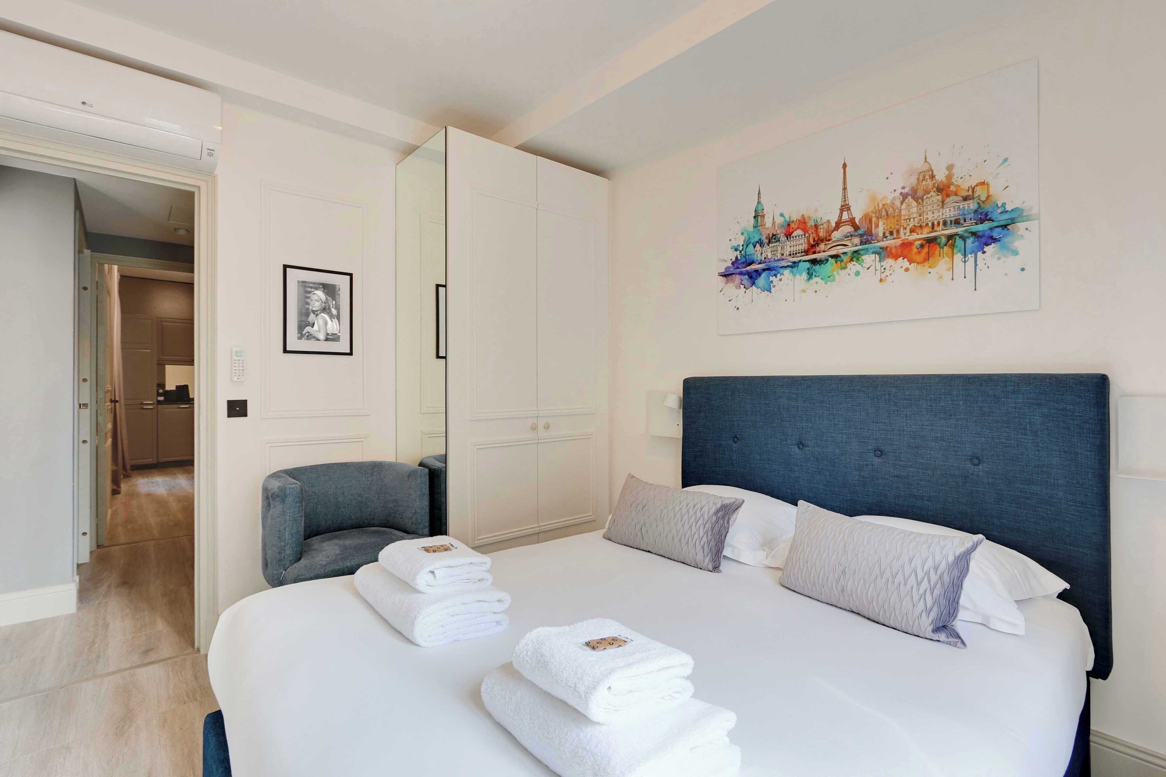 Hotellihuone, jossa on suuri sänky, vaatekaappi, tuoli ja seinällä kaupunkimaisemaa esittävä taulu.