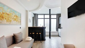 A modern hotel room with a sofa, a dining area with a bar, a flat-screen TV, and a large window. - Résidence Paris CHARONNE (Paris)