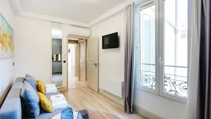 A modern living room with a sofa, a coffee table, and a TV. There is a window with curtains and a view of the outside. - Résidence Paris CHARONNE (Paris)