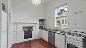 A kitchen with white cabinets, a gas stove, a dishwasher, a window with a view of a house, and a fireplace. - Hewlett Cottage - Central Pet Friendly (Port Fairy)