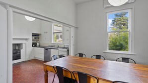 A modern kitchen with white cabinetry, a wooden dining table, and a large window offering a view of greenery. - Hewlett Cottage - Central Pet Friendly (Port Fairy)
