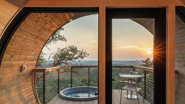 Une terrasse en bois avec un jacuzzi, une table et des chaises, offrant une vue panoramique sur un paysage magnifique, notamment au coucher du soleil.