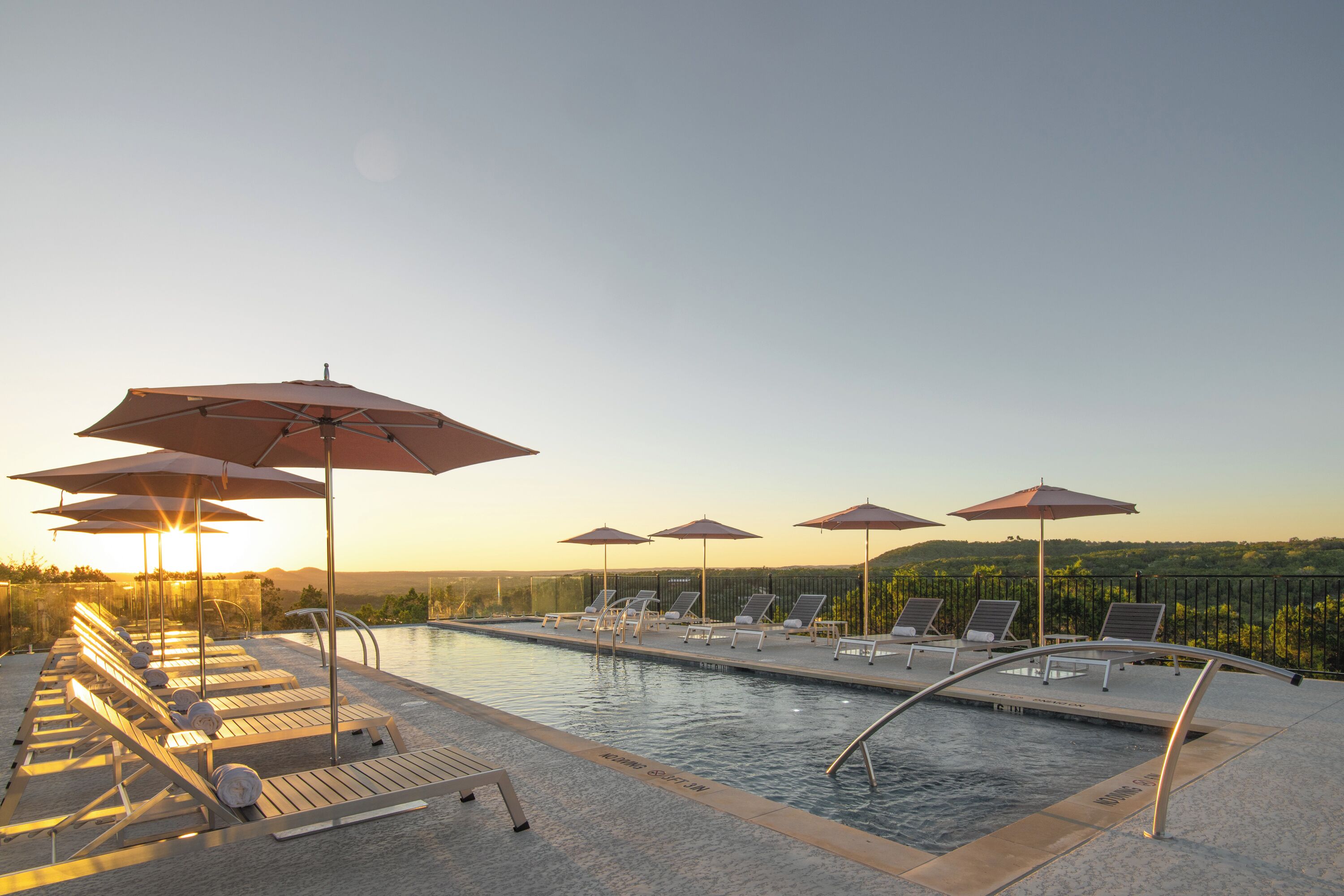 A poolside area with lounge chairs and umbrellas, a clear sky, and a view of the landscape in the background.