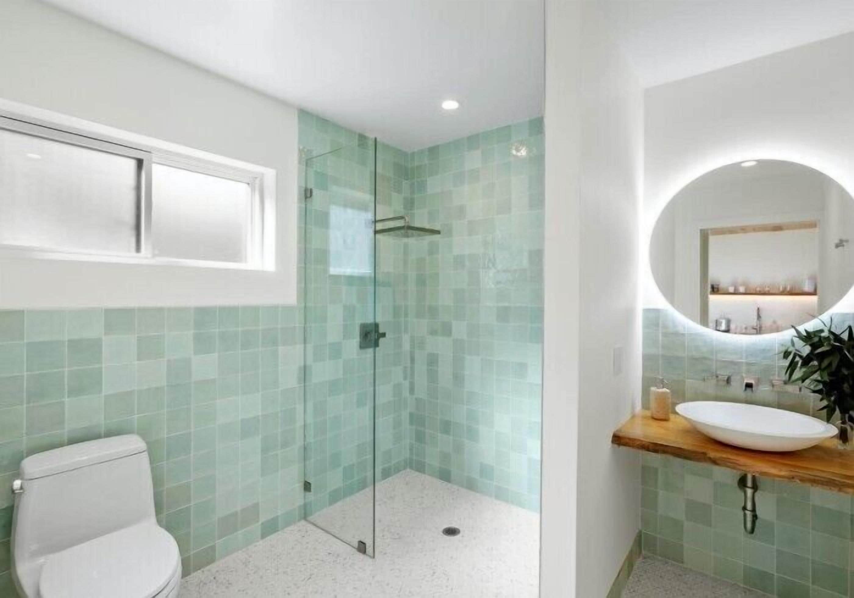 A bathroom with a glass-enclosed shower, a white toilet, a sink with a wooden countertop, and a mirror with a round frame.