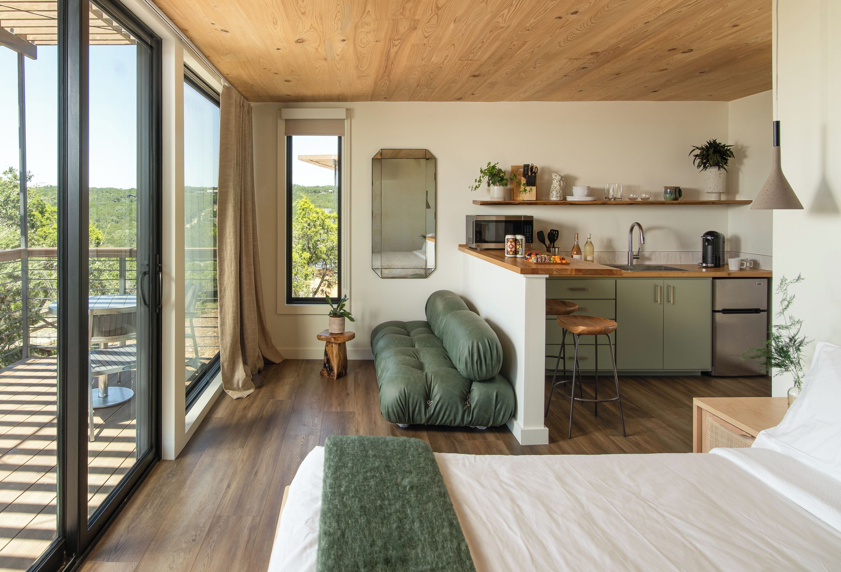 A modern room with a bed, a green sofa, a kitchen with a sink and a microwave, and a large window offering a view of greenery.