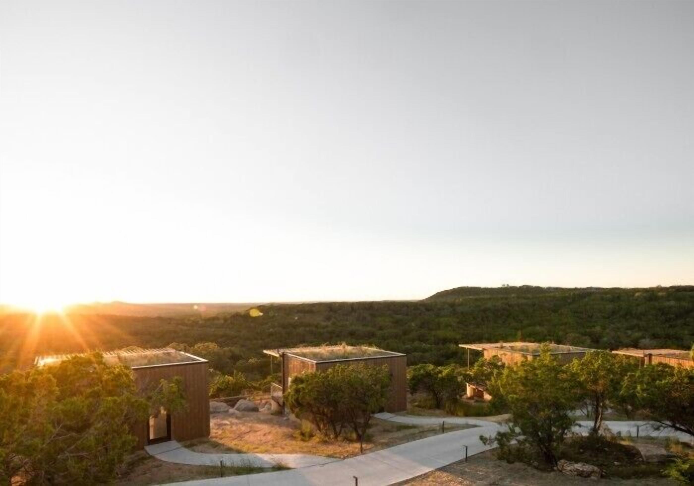 Un moderno complejo de edificios con varias estructuras, rodeado por un paisaje natural con árboles y colinas, durante el atardecer.