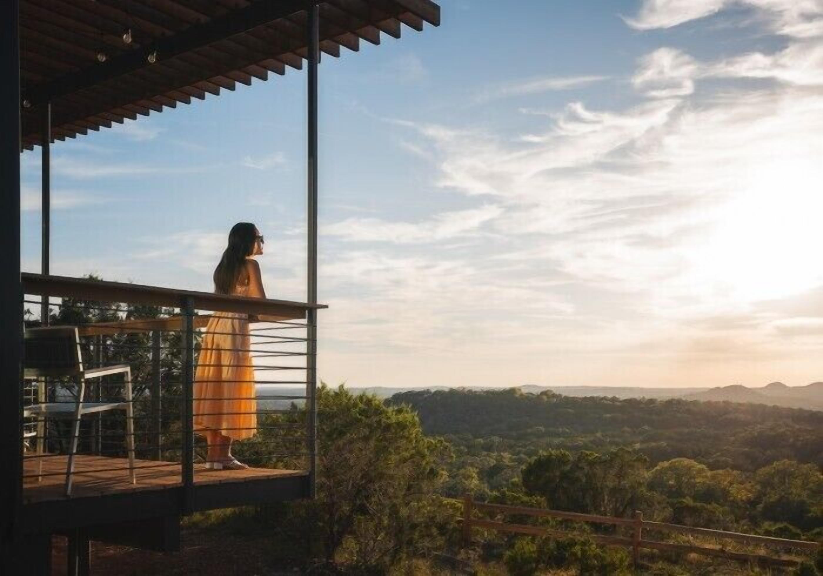 A person standing on a balcony overlooking a scenic landscape with trees and hills.