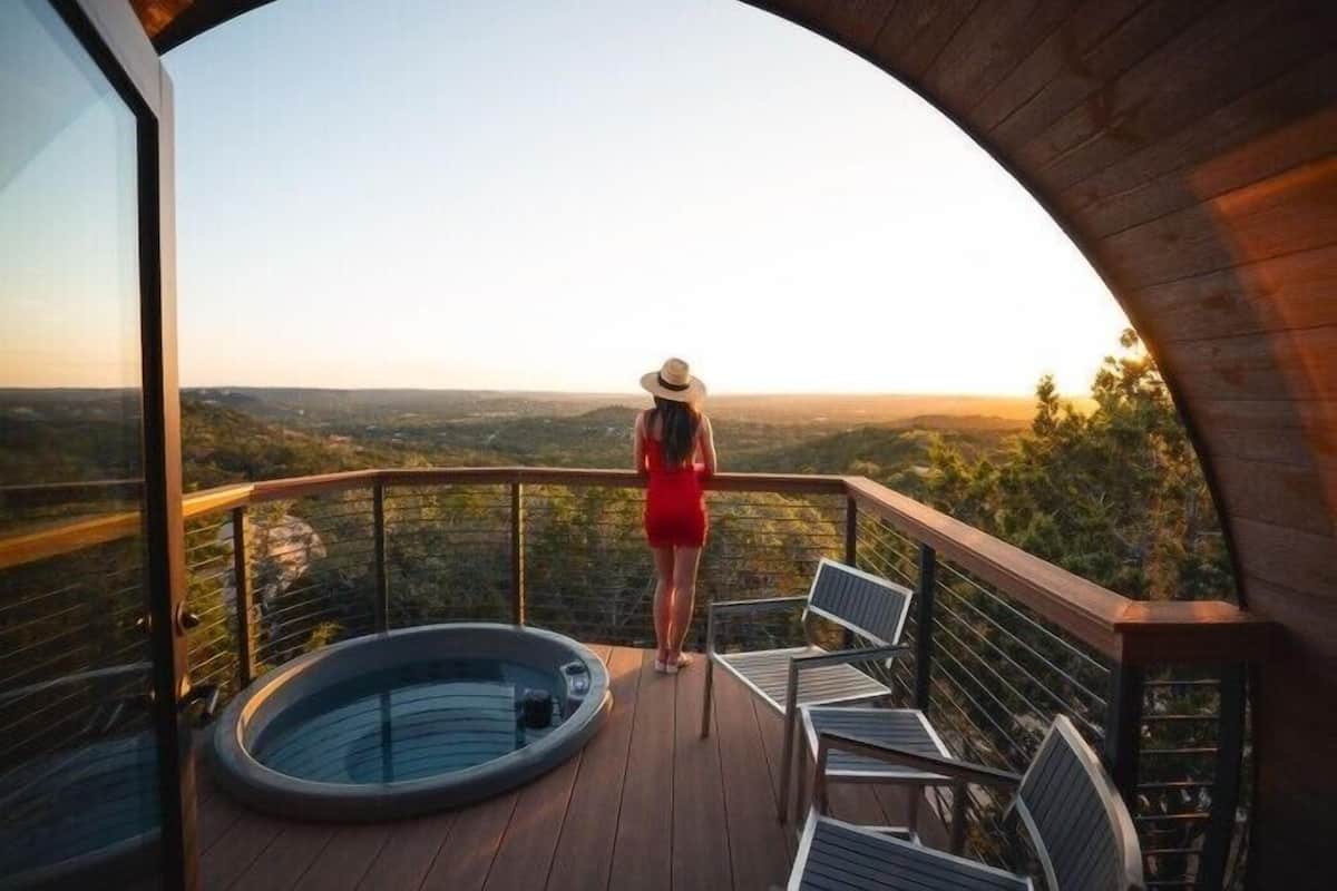 A woman in a red dress standing on a balcony with a hot tub and chairs, overlooking a scenic landscape.
