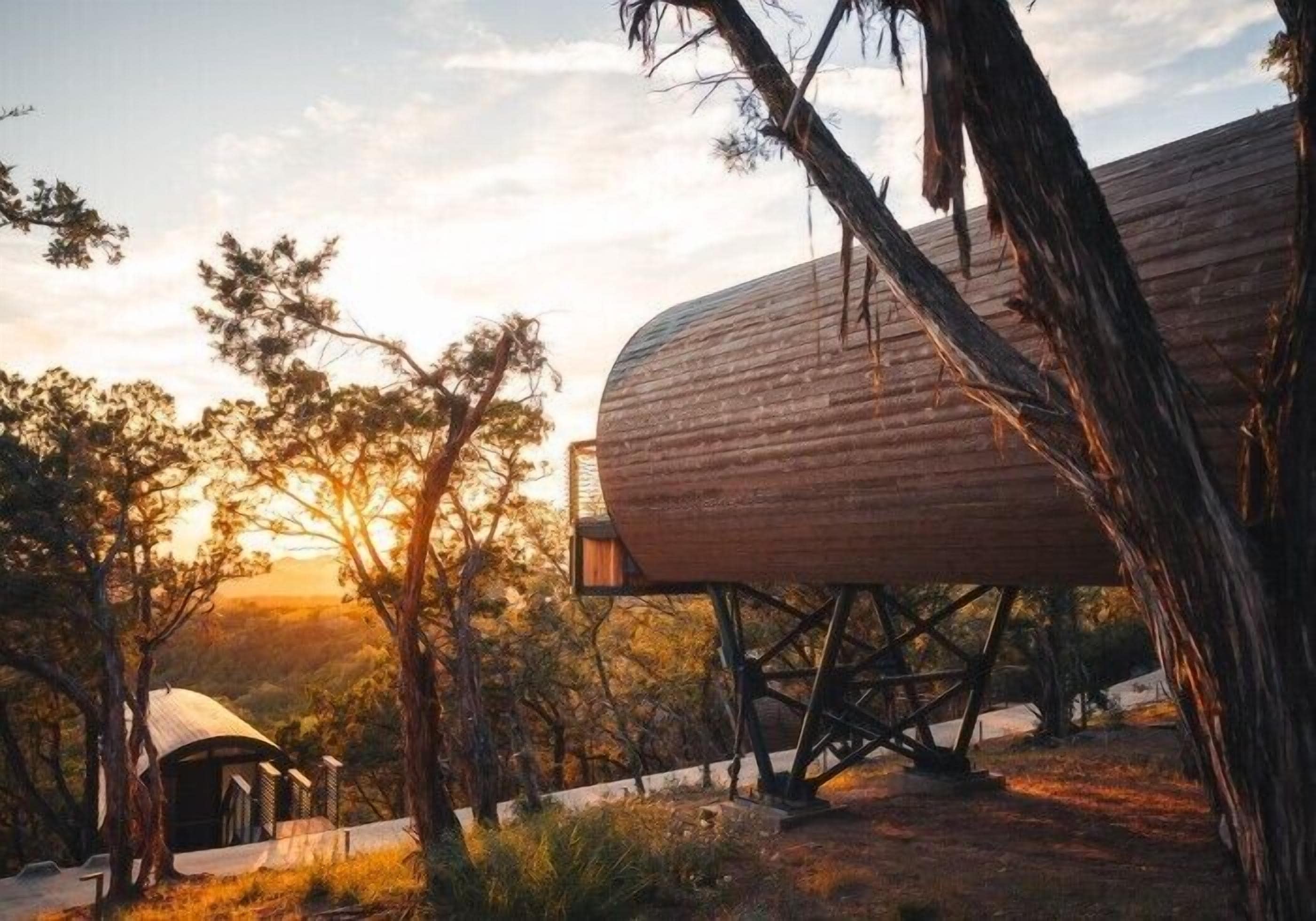 Una estructura rústica de madera con techo abovedado, elevada sobre pilotes, rodeada de árboles y un cielo despejado.