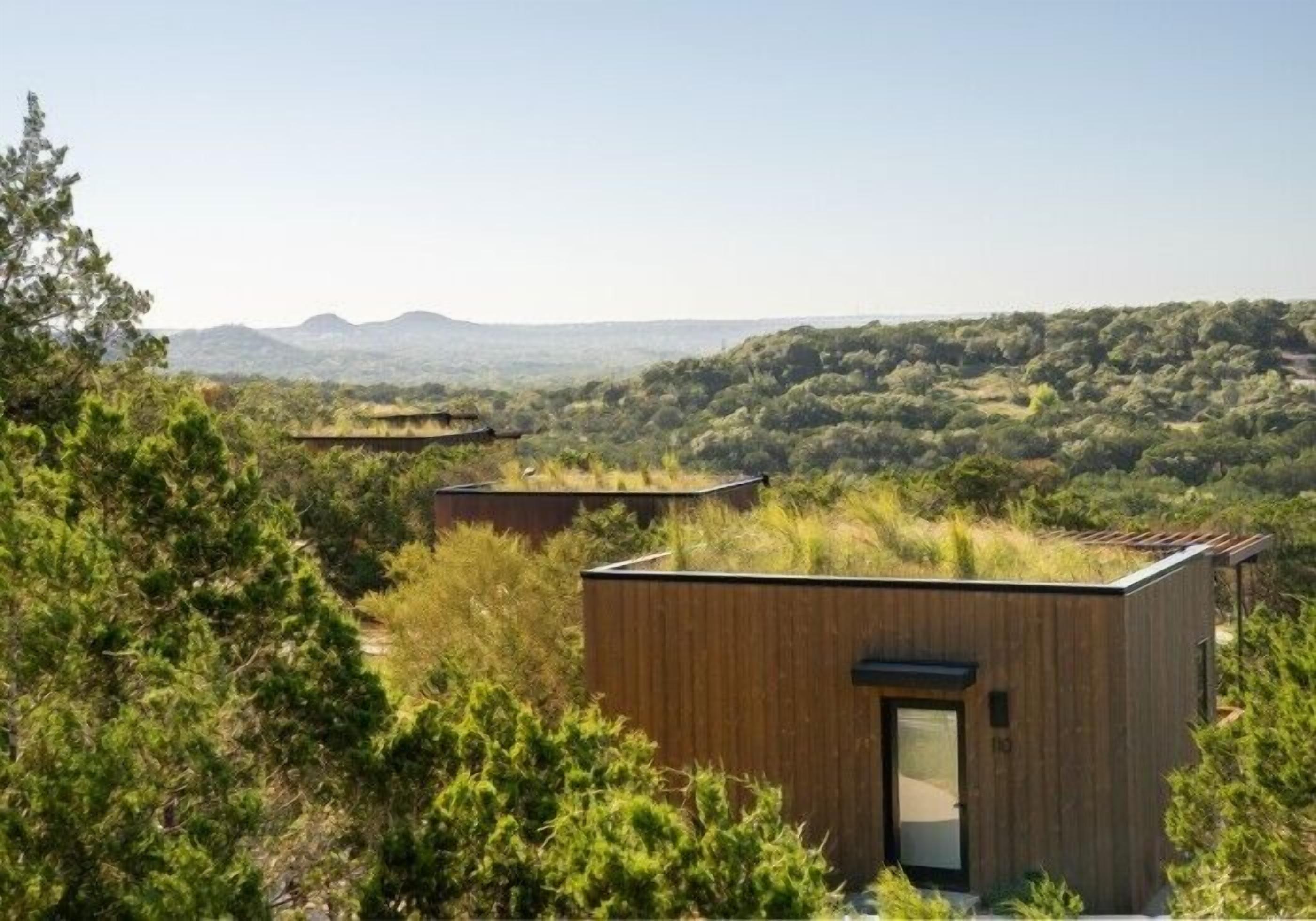 Une cabane moderne en bois, dotée d’un toit plat, entourée d’une végétation luxuriante et avec des collines en arrière-plan.