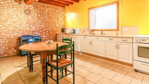 Una cocina con armarios blancos, una mesa de madera y sillas. Se observa una pared de ladrillo y una ventana sobre el fregadero.