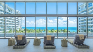 A room with large windows overlooking a beach and ocean, featuring lounge chairs and a small table. - Hotel Mousai Cancun Ocean Front Adults Only - All Inclusive (Punta Sam)