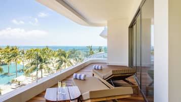 Ein Balkon mit Holzboden, zwei Liegestühle mit Kissen, ein kleiner Tisch mit einem Getränk und Blick auf Strand und Meer.