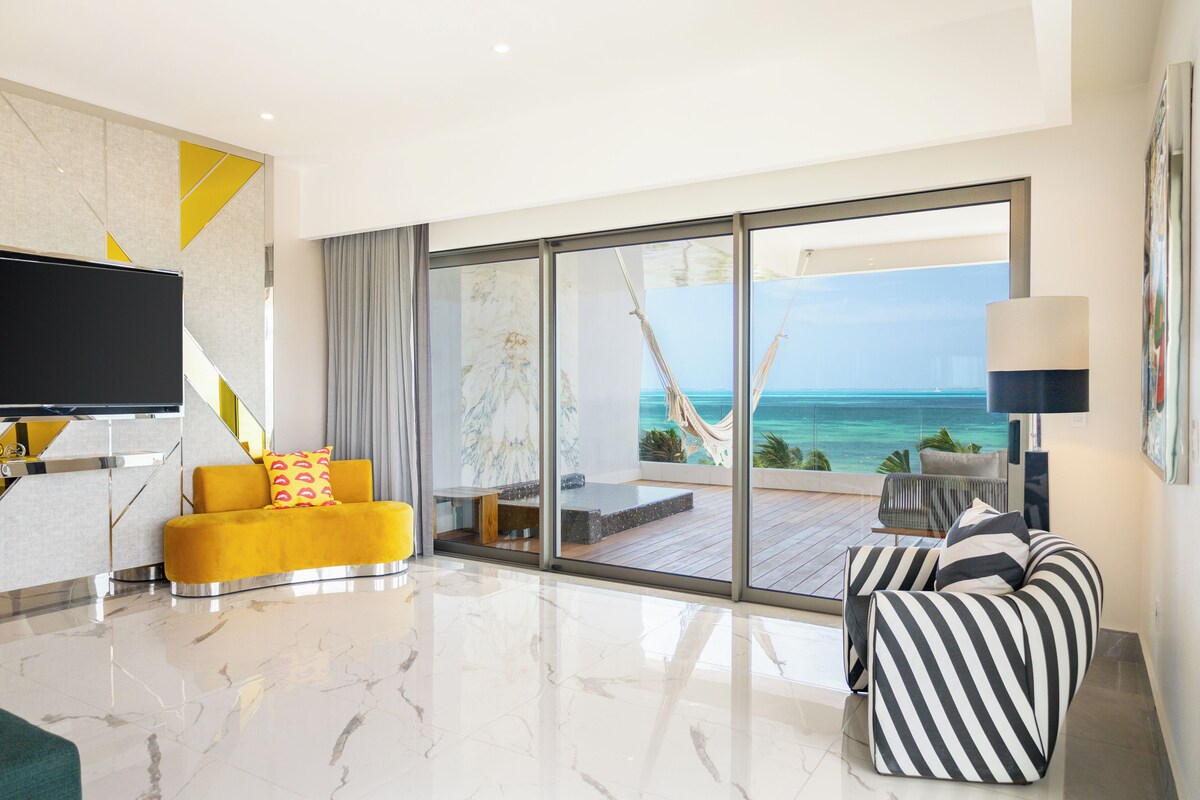 A modern living room with a large TV, yellow and black striped sofa, and a view of the ocean through floor-to-ceiling windows.
