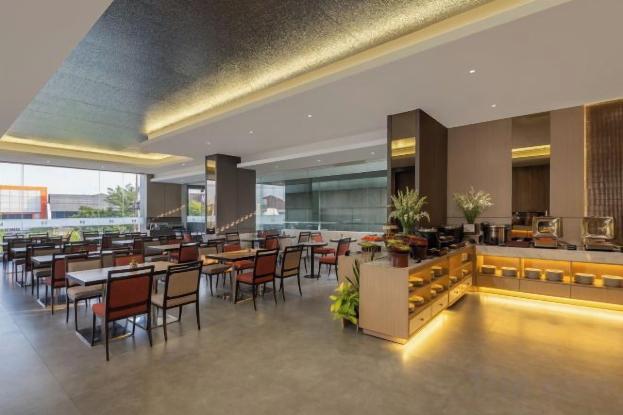A spacious hotel lobby with multiple dining tables and chairs, a reception desk, and large windows offering a view of the outdoors.