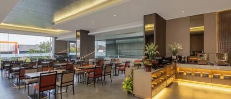 A spacious hotel lobby with multiple dining tables and chairs, a reception desk, and large windows offering a view of the outdoors.