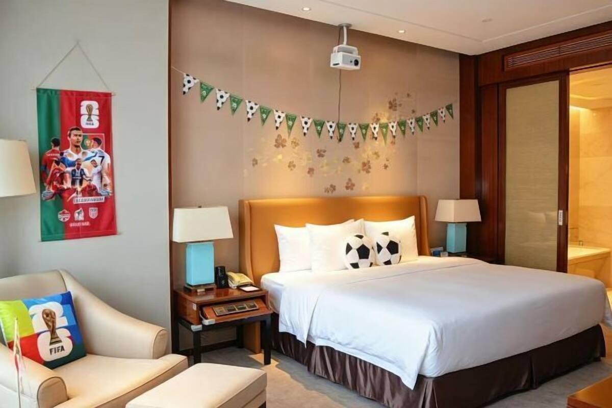 A hotel room with a bed, a desk, a chair, a nightstand, a lamp, a projector, and a wall decoration.