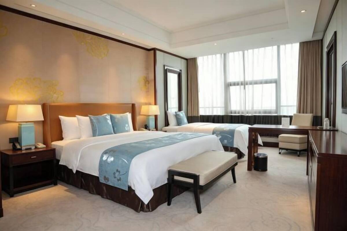 A hotel room with two beds, a desk, a chair, a bench, and a large window with curtains.