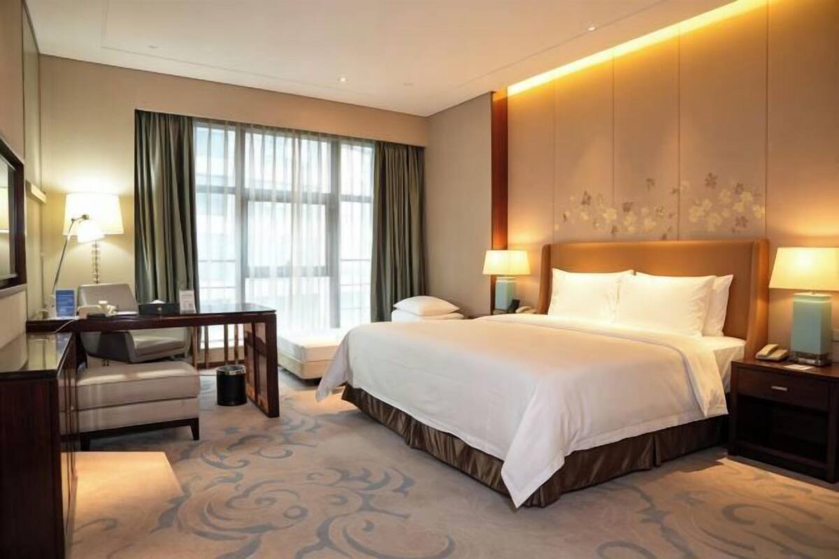 A hotel room with a bed, bedside tables, a desk, a chair, a lamp, and a large window with curtains.