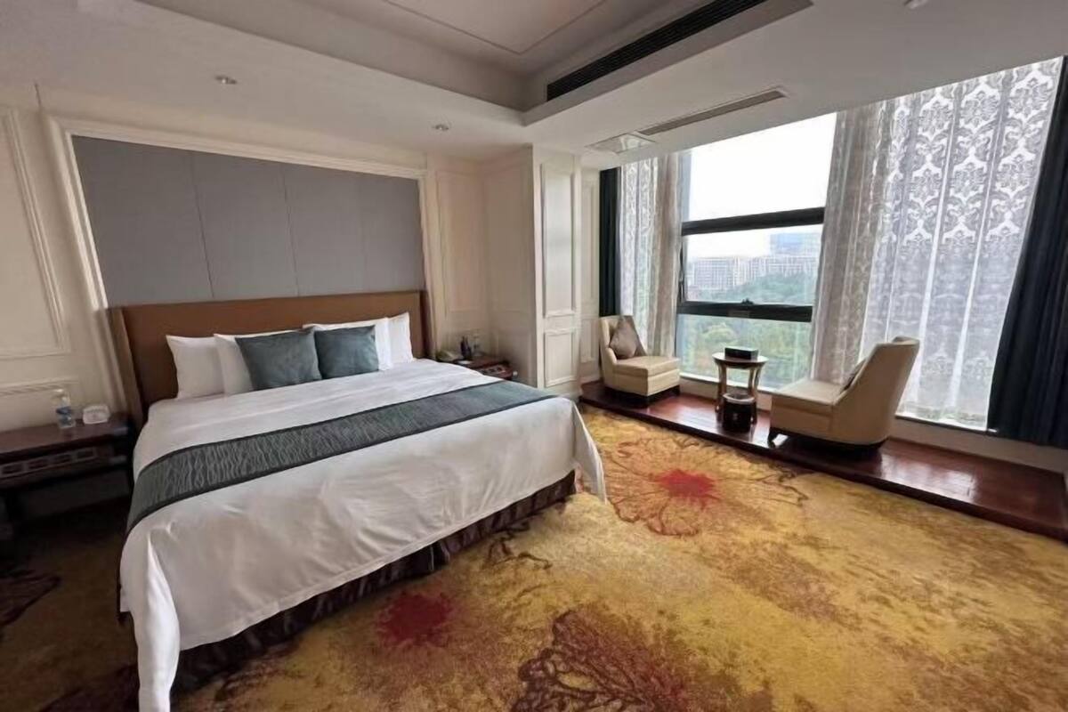 A hotel room with a large bed, a view of the city, a small table, and a chair by the window.
