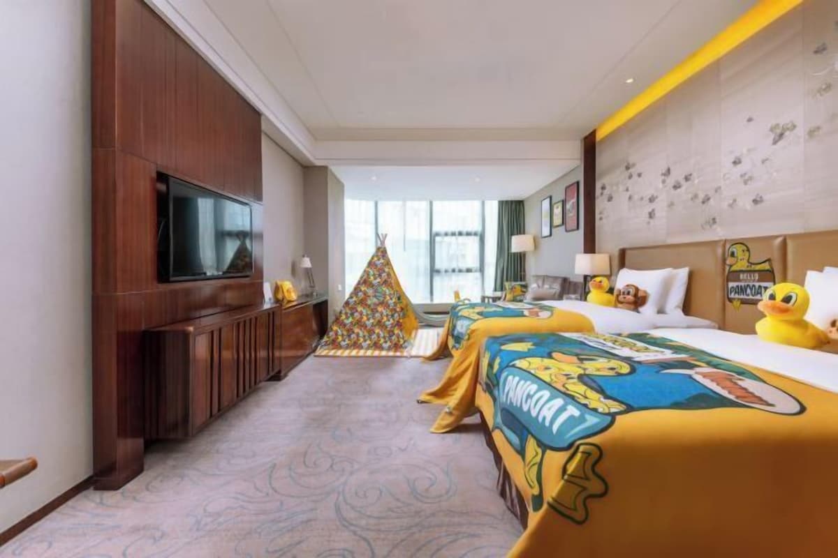 Hotelkamer met een groot flatscreen televisietoestel, een houten kast, een kleurrijke driehoekige decoratie, twee bedden met beddengoed met tekenfilms en een muur met decoraties van eenden.