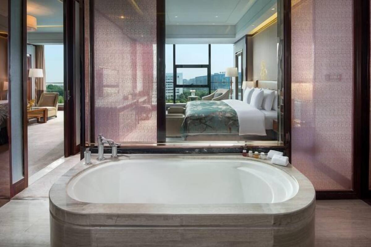 A modern hotel room with a large bathtub, a bed with white linens, and a view of the city through large windows.
