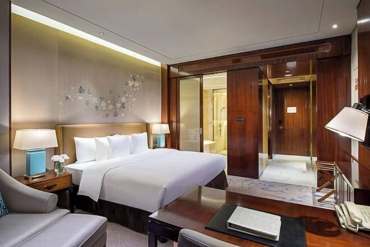 A hotel room with a large bed, bedside tables with lamps, a desk, and a chair. There is a wooden door leading to a bathroom.
