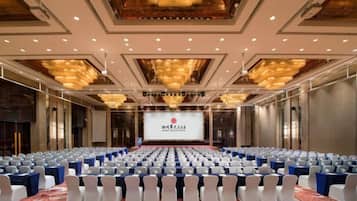 Un salón de conferencias con filas de sillas, un escenario con una pantalla y una araña.