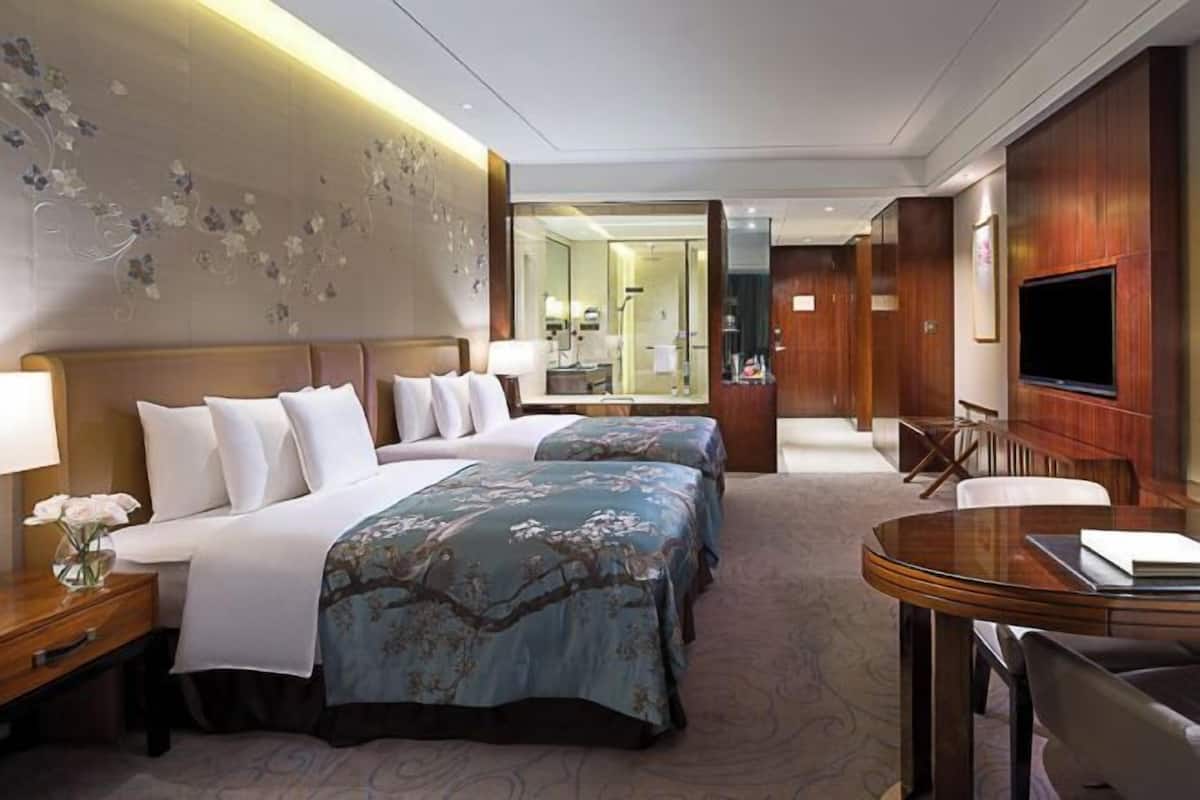 A hotel room with two beds, a desk, a chair, a TV, and a bathroom visible through a glass door.