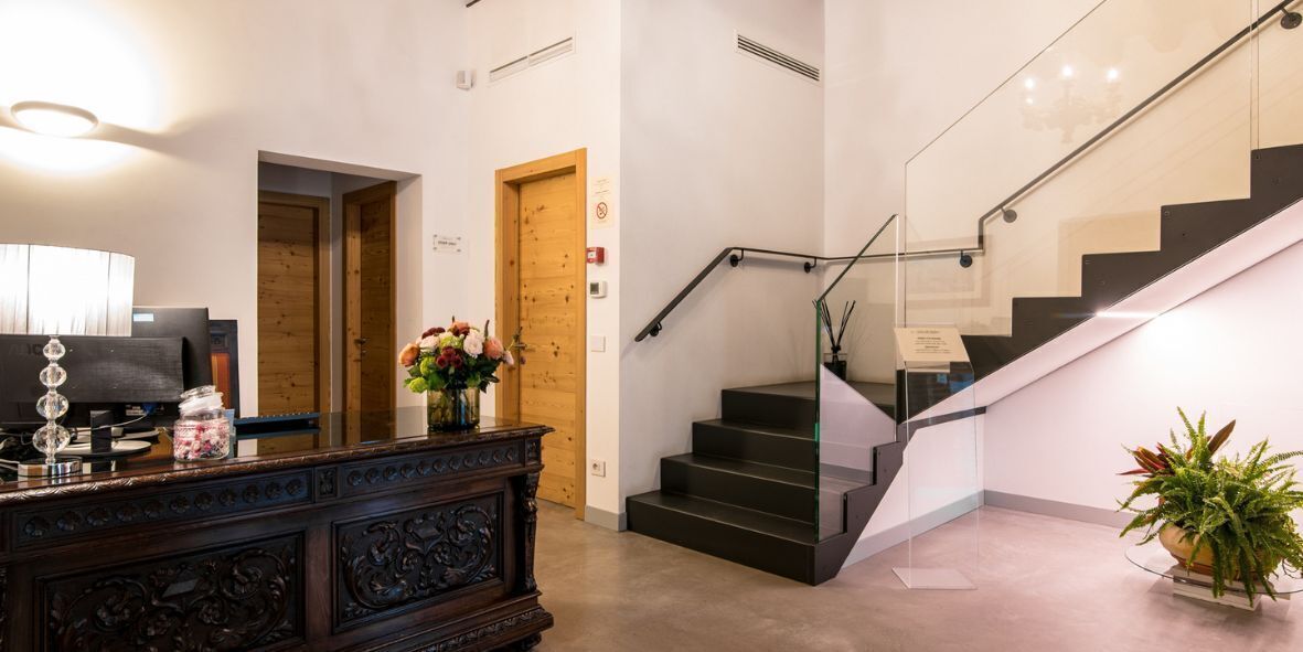 A modern hotel lobby with a wooden reception desk, a staircase with glass railings, and a potted plant.