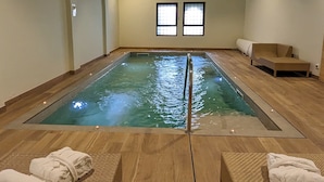 A spa room with a large indoor pool, wooden flooring, and two windows.