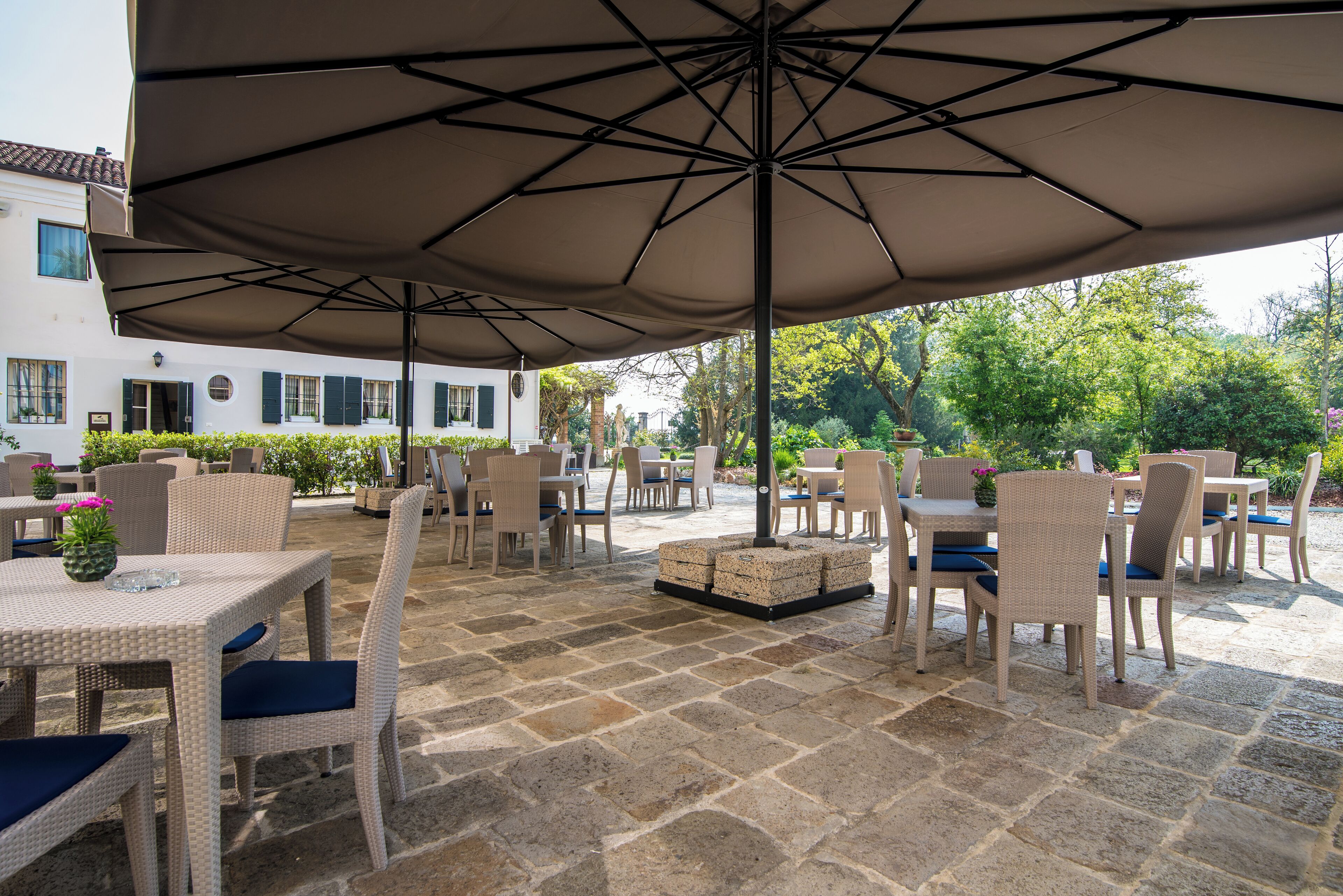 An outdoor patio with wicker furniture and umbrellas, stone flooring, and a view of a white building with shutters.