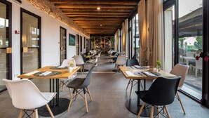 A modern restaurant with wooden beams and large windows. Tables are set with chairs, and the space is open and airy.