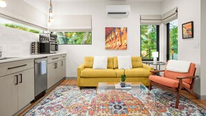 A modern kitchen with a coffee machine, microwave, and dishwasher. There is a yellow sofa with white cushions and a colorful rug. A small table with a plant is in front of the sofa. - Cozy studio, Pet Friendly, Outdoor Oasis near Zilker  (Austin)