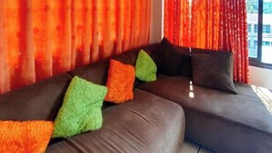 A modern living room with a sectional sofa, vibrant orange and green pillows, and red curtains. - RedDoorz Hostel @ Tita Aidz Inn Baguio (Baguio)