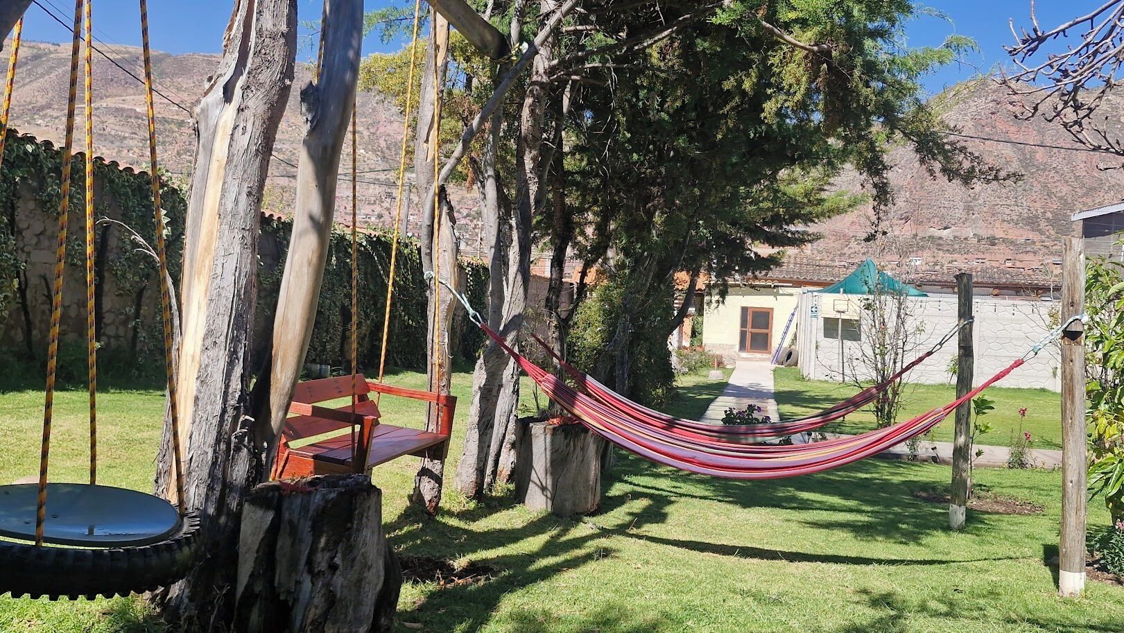 Una hamaca entre árboles, un conjunto de columpios y una casa al fondo.