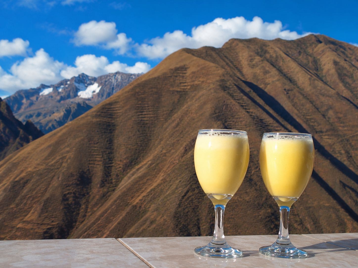 Dos vasos de una bebida amarilla sobre una superficie de mármol, con montañas al fondo.