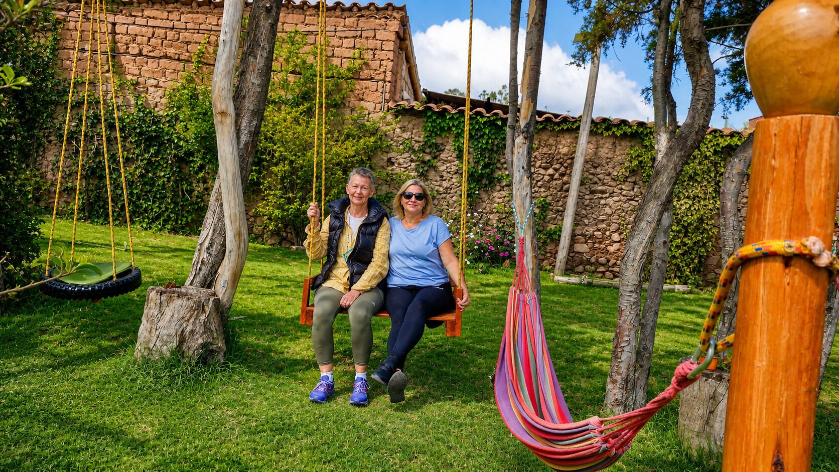 Two people on a swing in a grassy area with a stone wall and trees in the background.