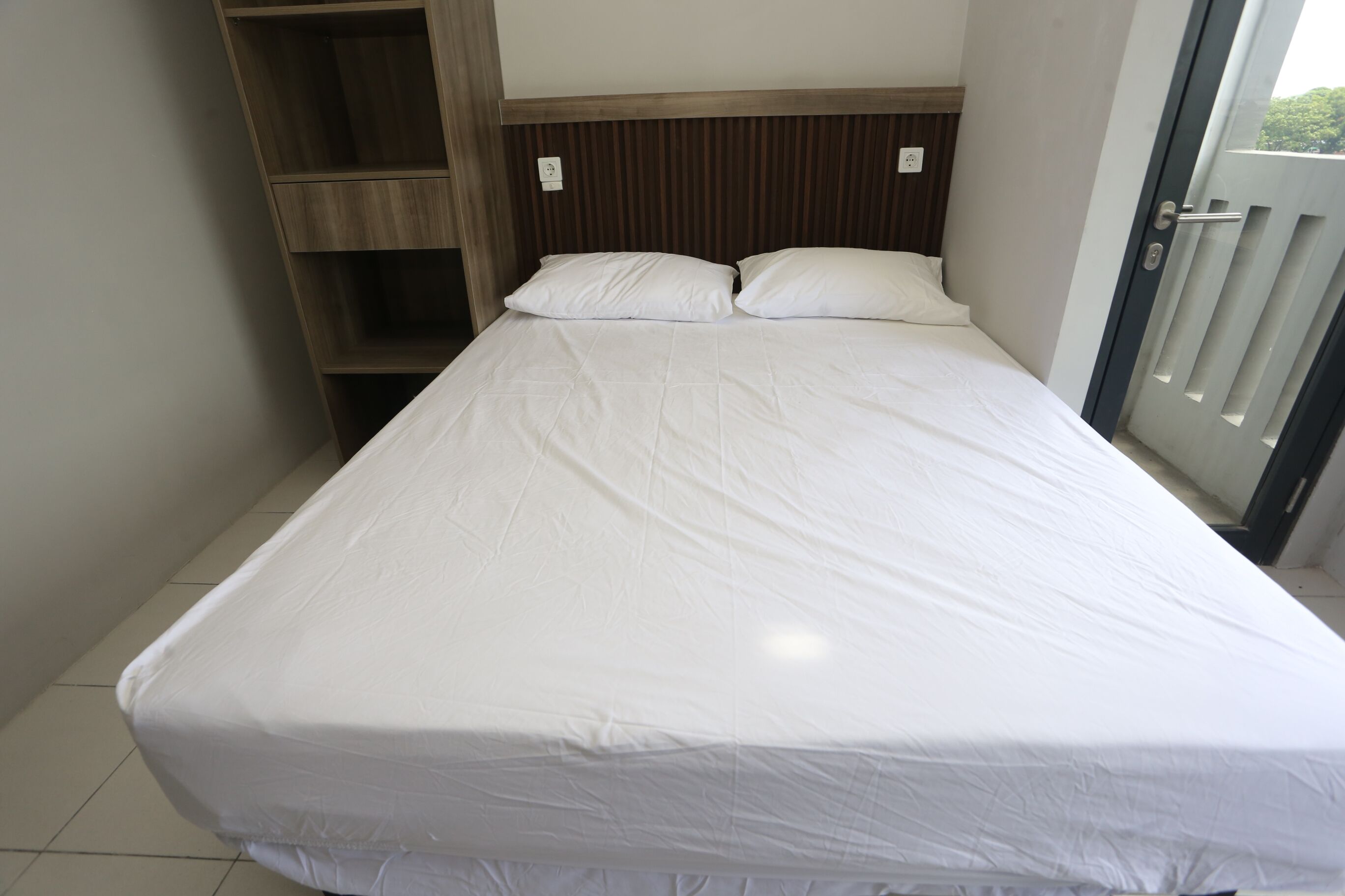 A single bed with a wooden headboard and white bedding in a room with a tiled floor and a door with a glass panel.