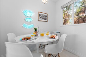 A dining area with a round table, white chairs, a neon eye decoration, a framed photo, and a window with a view of trees.