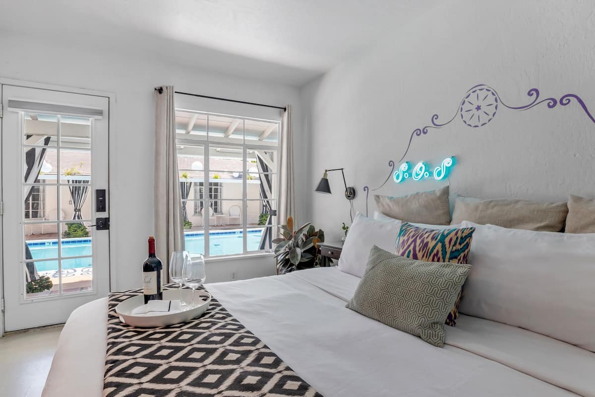 Ein Schlafzimmer mit einem Bett, einem Nachttisch mit einer Flasche und Gläsern, einem Neonzeichen an der Wand und Blick auf einen Pool durch ein Fenster.