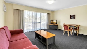 A hotel room with a red sofa, a wooden coffee table, a TV on a cabinet, a dining table with chairs, and large windows with curtains. - Suite with Kitchen, WiFi & Easy Access to Motorway (Mount Ommaney)