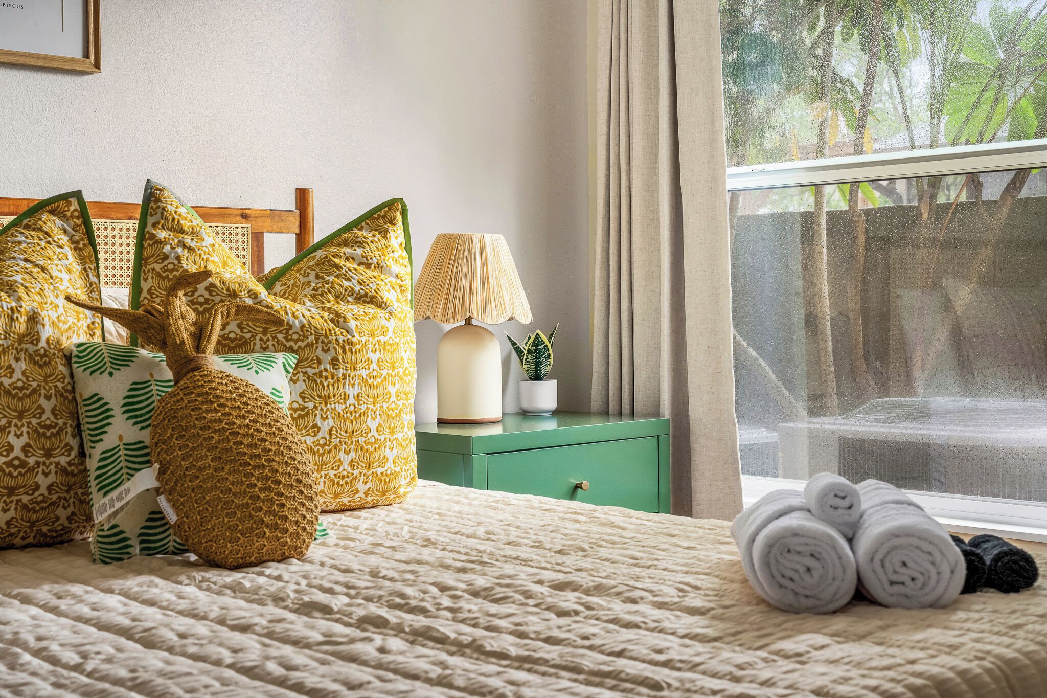 Ein ordentlich bezogenes Bett mit aufgerollten Handtüchern, einer Lampe und gemusterten Kissen auf einem grünen Nachttisch.