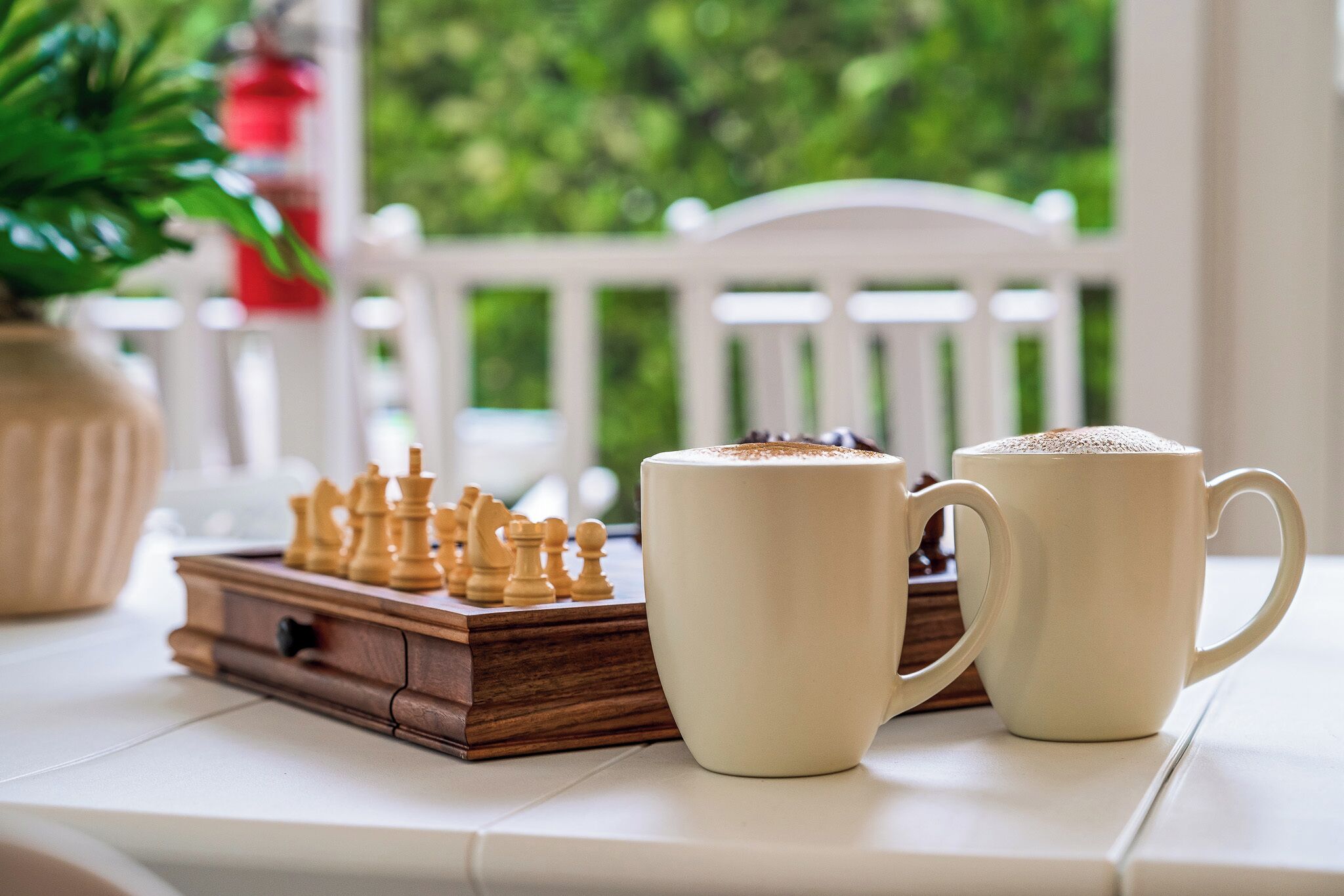 A chessboard and two coffee mugs on a table with a white chair in the background.