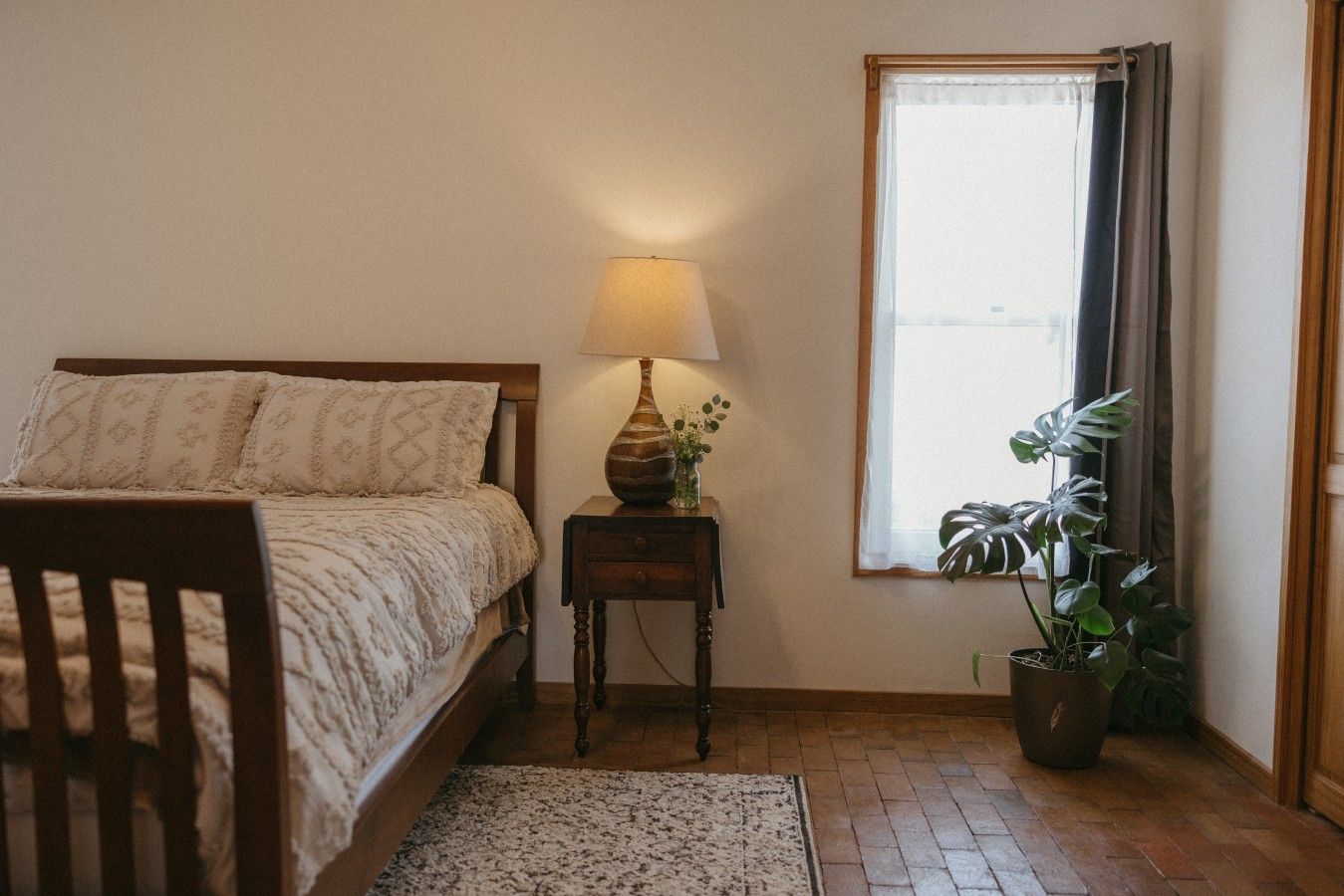 A bedroom with a bed, bedside table, lamp, potted plant, and a window with curtains.