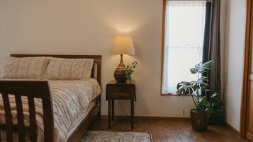 A bedroom with a bed, bedside table, lamp, potted plant, and a window with curtains.