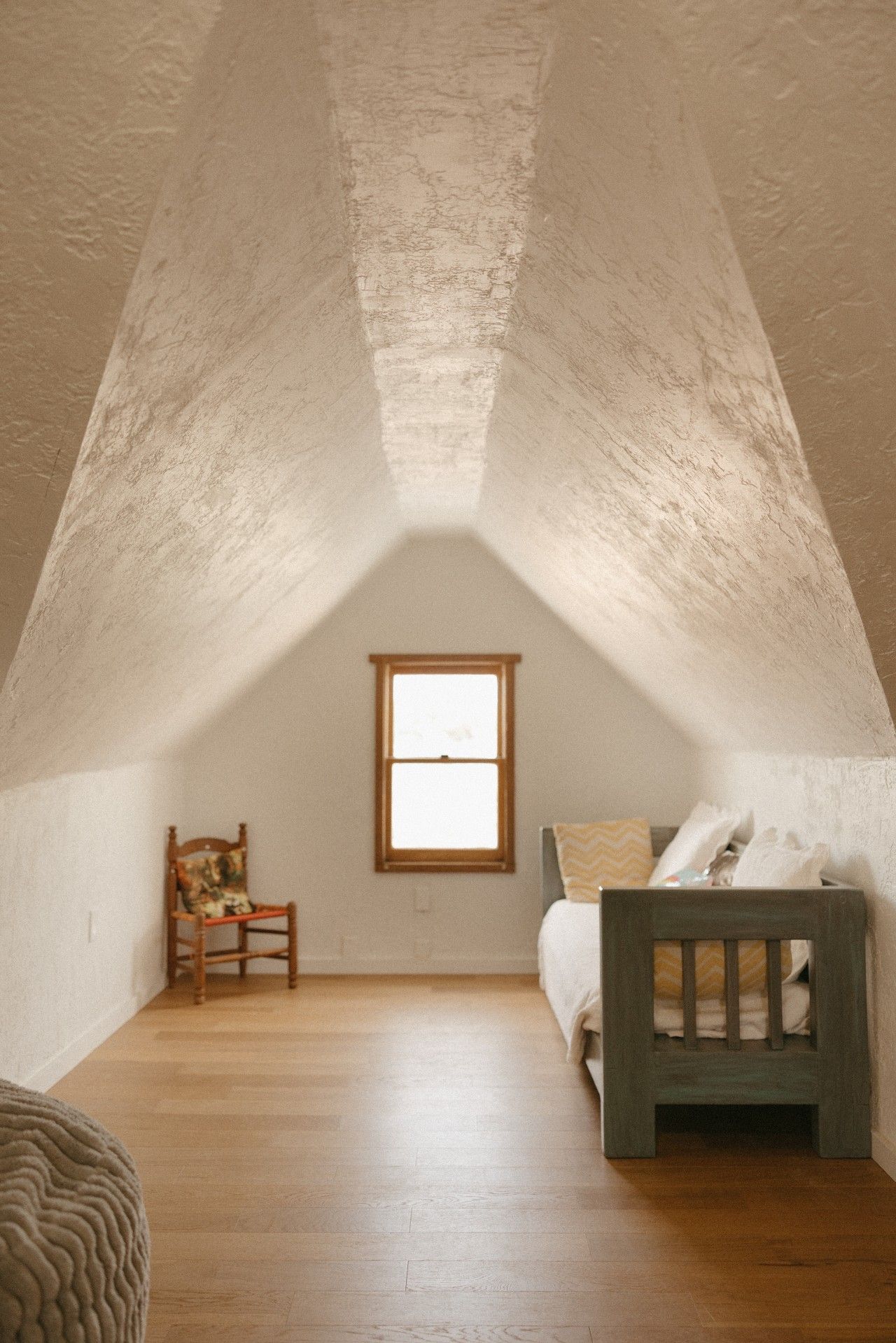 A small attic room with a sloped ceiling, a window, a wooden chair, a bed with a headboard, and a woven rug.