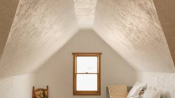 A small attic room with a sloped ceiling, a window, a wooden chair, a bed with a headboard, and a woven rug.