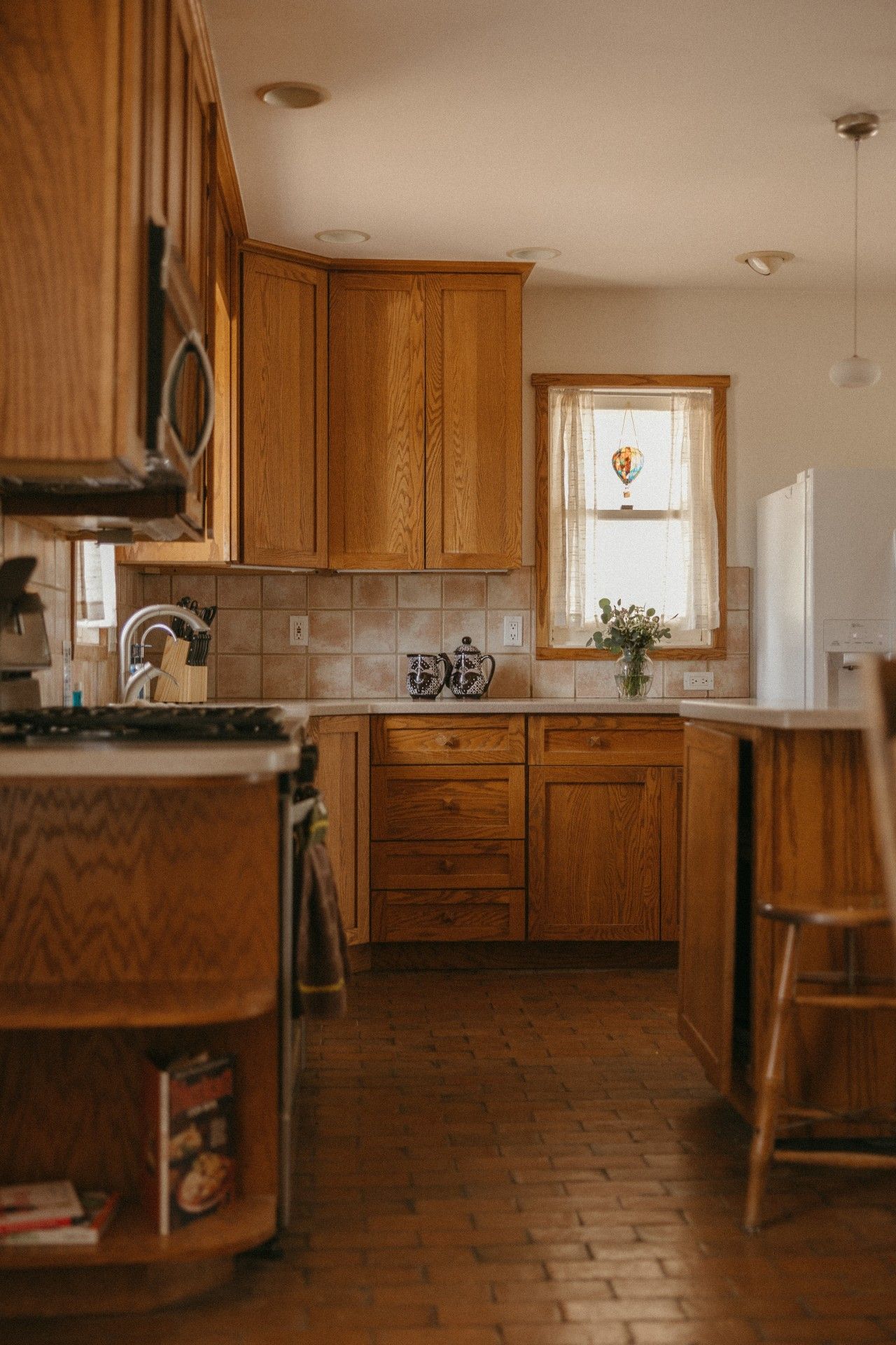 Une cuisine avec des meubles de rangement en bois, un pare-feu en carrelage, une fenêtre avec un rideau et un sol en briques.